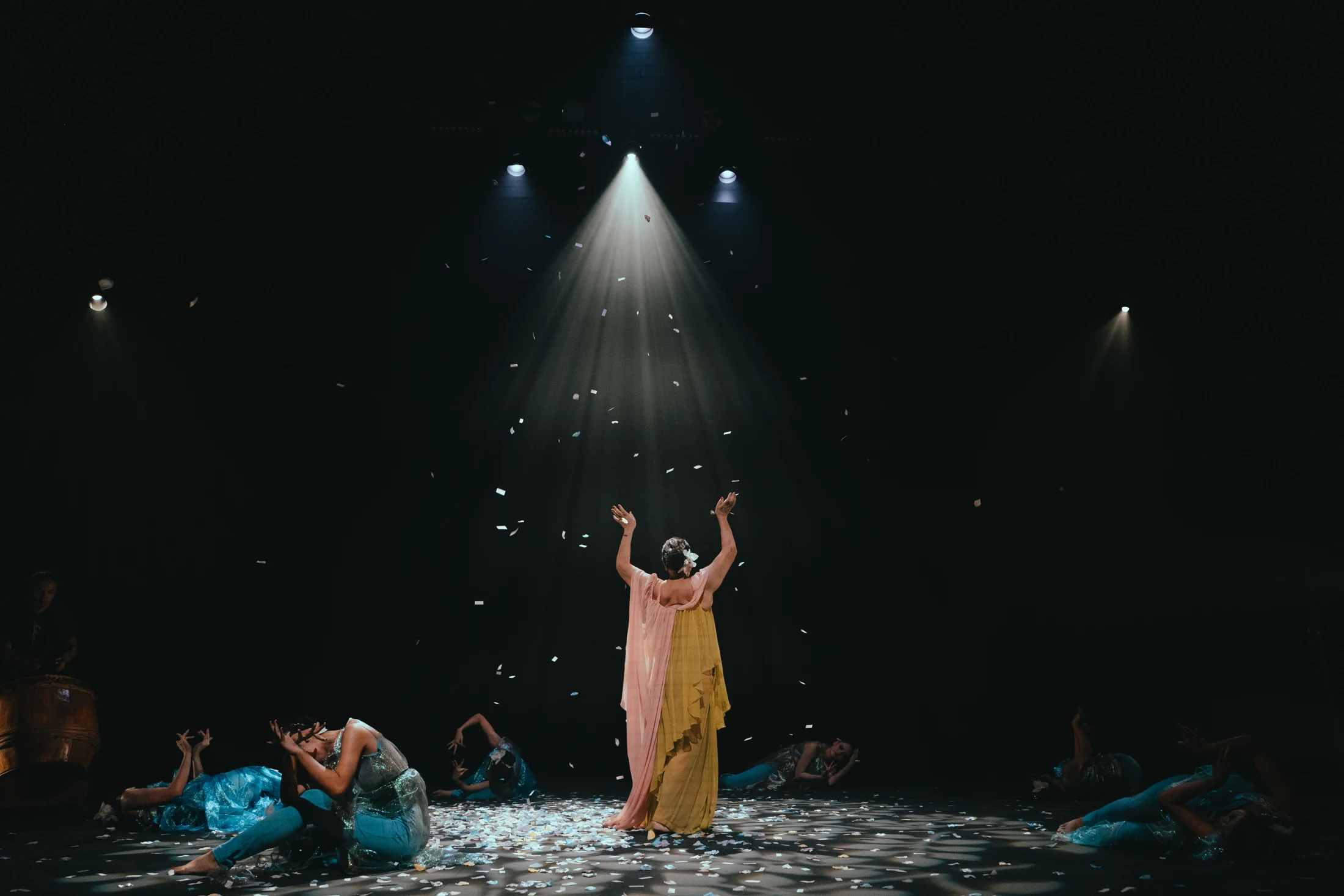 A Kelopak performer in yellow reaches upward as petals fall beneath a spotlight