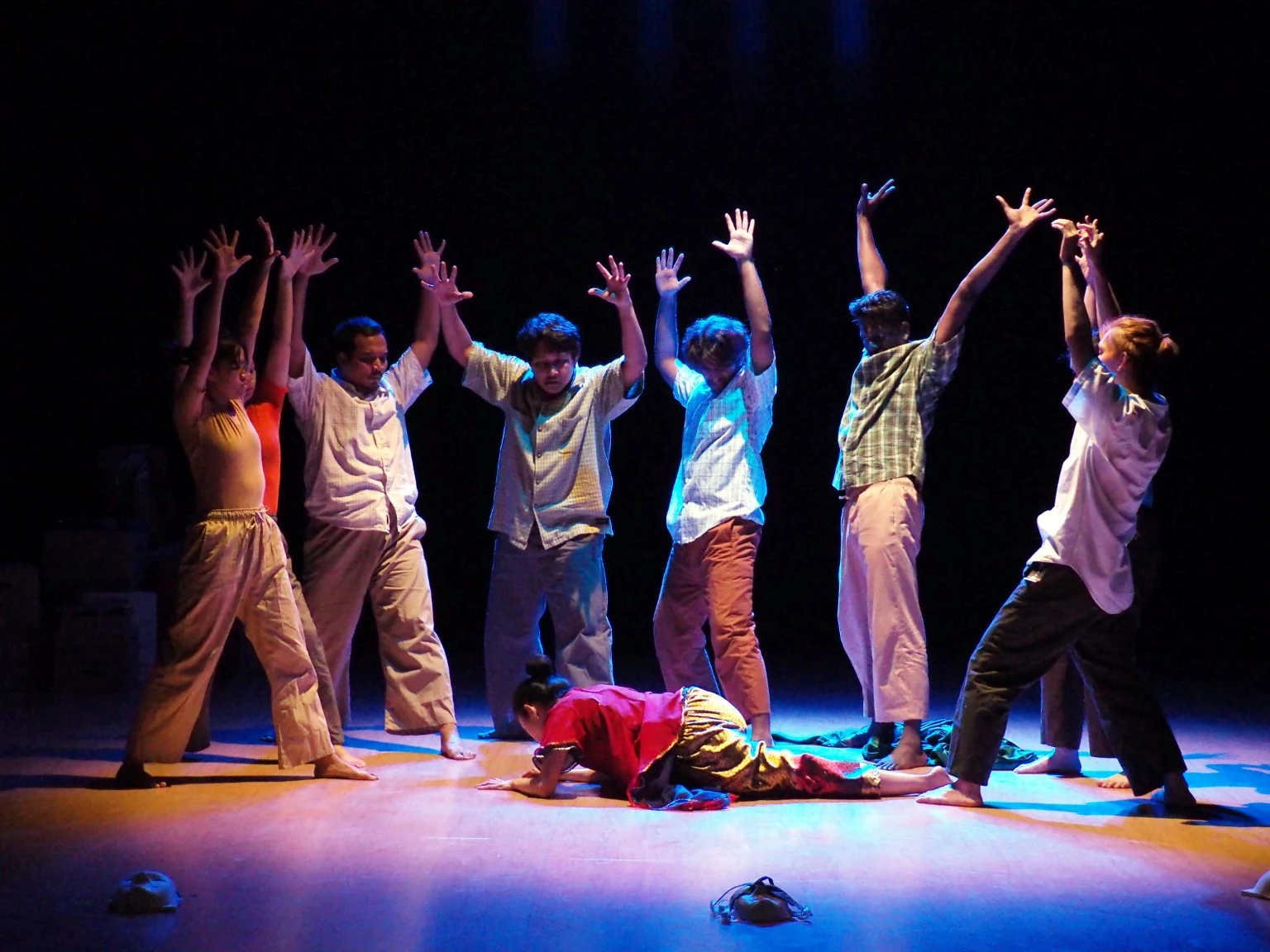 The Lari ensemble raises their arms around a performer on the floor under blue and amber stage lighting