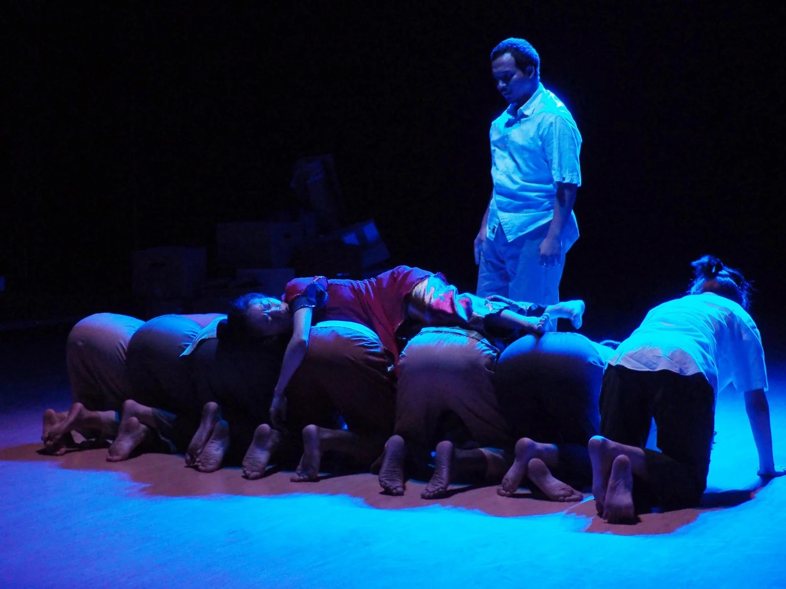 A Lari performer in white stands above an ensemble forming a low line across the stage
