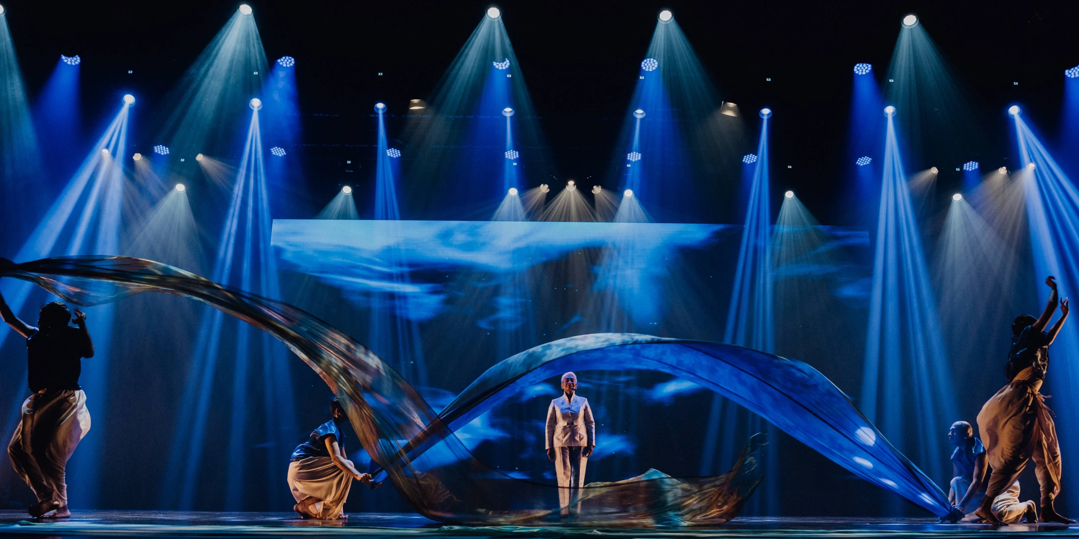 Performers move long blue fabric across a stage beneath beams of blue light
